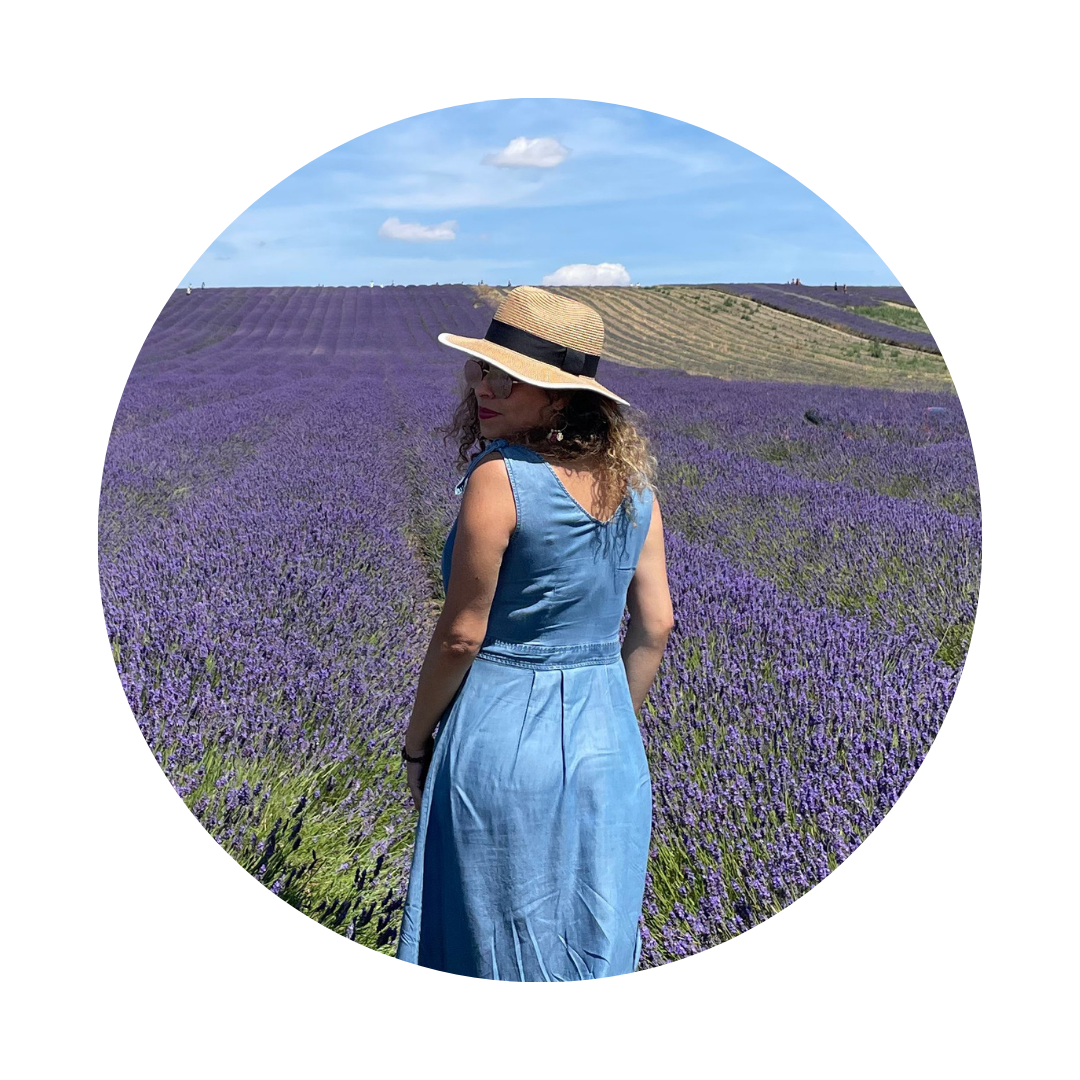 Woman in a sunlit lavender field in England, facing away from the camera, wearing a straw hat and sunglasses. The purple flowers stretch into the distance under a bright blue sky, evoking a peaceful and intentional solo moment.